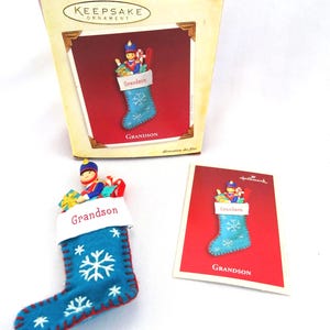 May include: A Hallmark Keepsake Christmas ornament in a box. The ornament is a blue felt stocking with white snowflake designs and the word "Grandson" on the cuff. A toy soldier and gifts are inside.