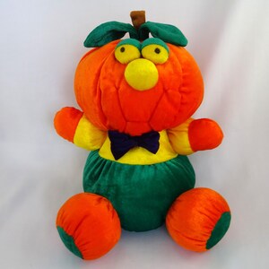 May include: A plush pumpkin character with an orange head, green leaves, and yellow eyes and nose. It wears a yellow and green outfit with a purple bow tie and orange feet. A whimsical Halloween or fall-themed toy.