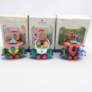 May include: Three Hallmark Easter train ornaments in their original boxes. Each train car is brightly colored with whimsical designs, including a basket of chicks, a teacup, and a bunny. The ornaments are on a white background.