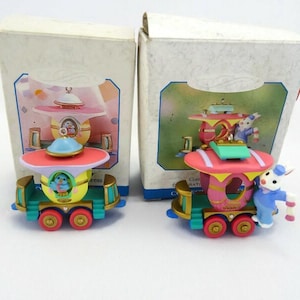 May include: Two decorative train ornaments with colorful designs. One train has a blue bird in the center, and the other has a bunny figure. The trains have pink, yellow, and teal accents, with red wheels. The ornaments are in their original boxes.