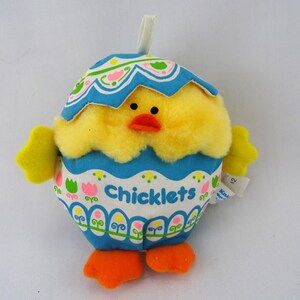 May include: A plush toy shaped like a yellow chick emerging from a blue and white patterned eggshell. The eggshell features the word Chicklets in blue text, decorated with pink and green tulip motifs and a white loop at the top for hanging.