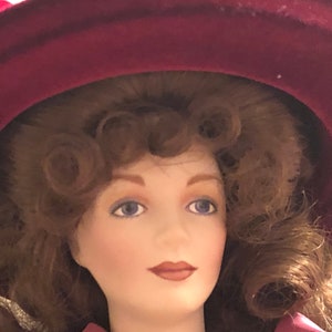 Pode incluir: Uma boneca de porcelana com um chapéu bordô e cabelo longo e encaracolado castanho. A boneca tem olhos azuis e está vestindo um vestido rosa.
