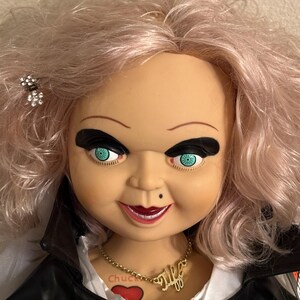May include: A doll with pink hair, black eye makeup, and teal eyes. The doll wears a black jacket and a gold necklace that says "Tiff". A small black beauty spot is visible on the doll's face.
