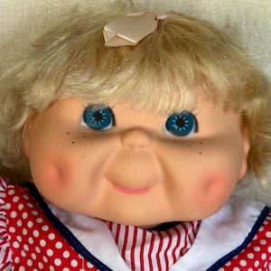 Puede incluir: Una muñeca vintage con cabello rubio y ojos azules. La muñeca tiene mejillas rosadas y pecas. Lleva un vestido de lunares rojos y blancos con cuello blanco y corpiño a rayas rojas y blancas. Un pequeño lazo está en el pelo de la muñeca.