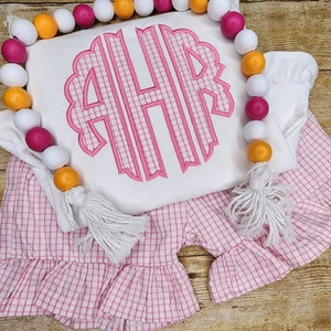 May include: White top with a pink monogram and matching pink gingham shorts. The monogram features the letters AHR in a scalloped design. A beaded garland with white, pink, and orange beads is draped across the outfit.