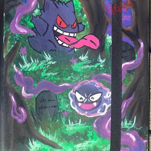 May include: A rectangular notebook featuring a hand-painted scene of a purple Gengar and a Gastly floating in a dark forest with green grass and gray tombstones. The cover includes swirling purple smoke, a black elastic closure, and red text.