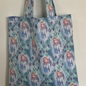 May include: A blue tote bag with a repeating pattern of a cartoon image of Elsa from Frozen. The pattern also includes a geometric design of diamonds and swirls.