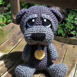 Op de afbeelding: Een gehaakt grijs en zwart hondenspeelgoed met een gele bloemenbedel. De hond heeft een droevige uitdrukking met grote ogen en een kleine zwarte neus.