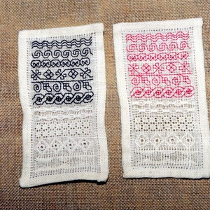 May include: Two white fabric squares with embroidered designs. The square on the left has a black embroidered design, and the square on the right has a pink embroidered design.