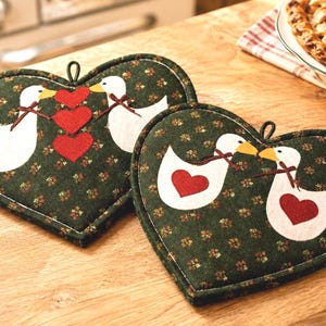 May include: Two heart-shaped pot holders made of dark green fabric with a small floral pattern. Each pot holder features white geese with yellow beaks and red heart appliqués. The geese are accented with small brown bows around their necks.