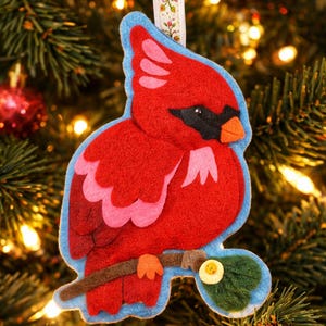 May include: A felt cardinal bird ornament featuring vibrant red plumage, pink wing accents, a black face mask, and an orange beak. The bird is perched on a brown branch with a green leaf and a small yellow button, suspended by a floral ribbon.