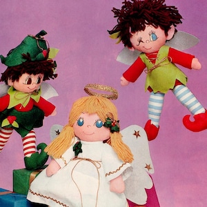 May include: Three handmade fabric dolls: an angel with blonde hair and a halo, and two elves with red and green outfits. The angel wears a white dress, and the elves have striped legs. All dolls have wings and are set against a pink background.