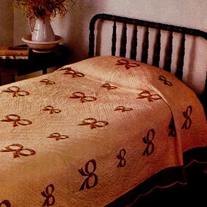 Op de afbeelding: Een vintage bed met een crèmekleurig sprei met een herhalend patroon van donkerbruine strikken. Het sprei heeft een geschulpte rand en ligt op een donkerbruin metalen bedframe. Een klein tafeltje met een vaas en boeken staat op de achtergrond.