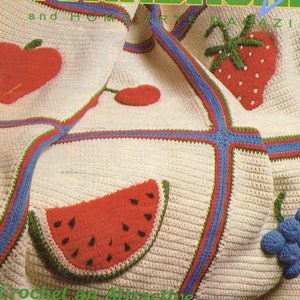 May include: A cream-colored crochet blanket featuring colorful fruit appliques including a red apple, a red strawberry with white seeds, a red watermelon slice with black seeds, and a cluster of blue grapes, all bordered by blue and green yarn.