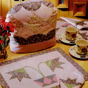 Puede incluir: Un juego de té acolchado que incluye un calentador de té, un mantel individual y una servilleta. El calentador de té es de color rosa claro con estampados florales y geométricos. El mantel individual presenta un diseño floral sobre un fondo rosa y un borde floral.