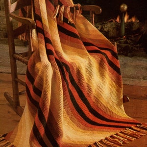 Op de afbeelding: Een gestreept gehaakt deken in tinten oranje, geel en zwart met franjes aan de randen. Het deken is gedrapeerd over een houten schommelstoel voor een open haard.
