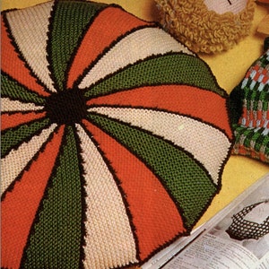 Może przedstawiać: Zbliżenie na kolorową, dzianą poduszkę z spiralnym wzorem w odcieniach zieleni, pomarańczy, bieli i brązu. Poduszka leży na żółtej powierzchni obok książki i okularów.