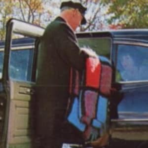 Puede incluir: Un coche negro vintage con un hombre con traje oscuro y sombrero abriendo la puerta. El coche tiene una ventana trasera y una puerta abierta. El hombre sostiene una manta colorida.