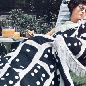 May include: A black and white crochet throw blanket with a domino pattern and white fringe. The blanket is draped over a chair with a person sitting on it.