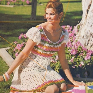 May include: A woman wearing a white crochet dress with colourful stripes and a floral pattern. She is sitting on a colourful blanket with a floral design.