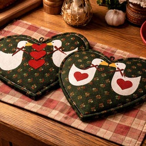 May include: Heart-shaped kitchen potholders with a green floral pattern and white ducks with red hearts. The potholders are on a red and white checkered cloth, with bread and other kitchen items in the background.