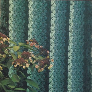 May include: A crocheted blanket with a green and blue ombre pattern. The blanket is made with a variety of stitches, including shells and stripes. The blanket is draped over a green bush with white flowers.