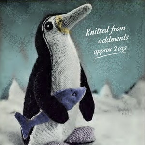 以下が含まれることがあります： 青い魚を持ったニットのペンギンのおもちゃ。ペンギンは白く、頭と羽根は黒いです。おもちゃは白い表面に置かれ、背景には青い水たまりがあります。テキスト「Knitted from oddments approx 2 ozs」は、青い背景に白で書かれています。