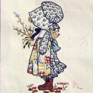 Puede incluir: Obra de arte enmarcada que representa a una niña con un gorro y un vestido de retazos, sosteniendo un ramo de flores azules. La obra está en un marco de madera y tiene una estética vintage. La niña lleva botas marrones y está de pie sobre hierba.