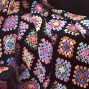 Op de afbeelding: Een handgehaakte deken met een patroon van veelkleurige granny squares verbonden door zwart garen. De vierkanten bevatten verschillende tinten roze, blauw, geel, oranje en groen. Een kleine zwarte voetenbank met een vlinderontwerp is aanwezig.