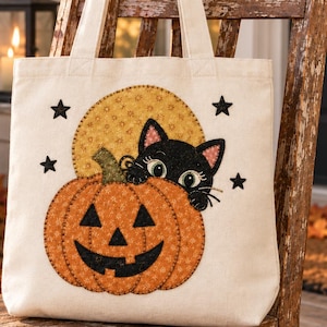 May include: A cream-colored canvas tote bag featuring an appliqued design of an orange jack-o'-lantern with a black cat peeking out from behind it. The design includes a yellow circular background, four black stars, and visible stitching details.