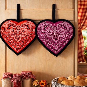 May include: Two heart-shaped pot holders with black trim, one red and one purple, hang on a wooden cabinet. A basket of rolls, jars, and a small flower arrangement sit on a red and white checkered tablecloth in a kitchen setting.