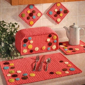 May include: A coral-colored crochet kitchen set featuring a bread box, placemat, trivet, and two pot holders. Each item is adorned with colorful, circular accents in yellow, brown, blue, and cream. A silver teapot and silverware are also visible.