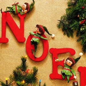 May include: Four small elf figurines dressed in green and red outfits are positioned on large, red, wooden letters spelling NOEL. The elves have pointed hats with white pom-poms and are interacting with the letters, which are set against a textured tan background with evergreen branches and a small Christmas tree.
