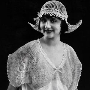 May include: A vintage black and white photograph of a woman wearing a crocheted cloche hat and a matching lace-trimmed top. The hat has a wide brim and is adorned with delicate floral accents. The top features a V-neck and a soft, flowing design.