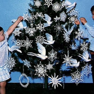 May include: Two children decorate a Christmas tree with white crocheted snowflakes and doves. The tree is decorated with white crocheted ornaments, including stars, snowflakes, and doves.