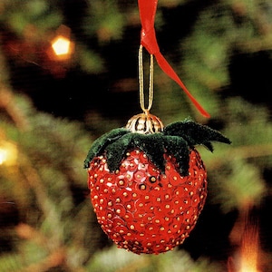 May include: A red Christmas ornament shaped like a strawberry, covered in sequins, with green leaves and a gold cap. It hangs from a red ribbon against a blurred background of a Christmas tree.