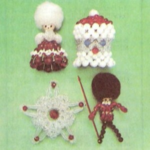Op de afbeelding: Vier handgemaakte kralenornamenten op een groene achtergrond. De ornamenten omvatten een pop met een witte, donzige kop en een rood-witte jurk, een kerstmanfiguur, een ster en een soldaat met een speer.