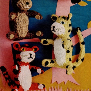 May include: A handmade knitted scene featuring three animal figures: a brown bear, a yellow leopard with black spots, and a red and black tiger. The figures are positioned against a backdrop of pink trees and a blue sky, creating a whimsical, handcrafted aesthetic.