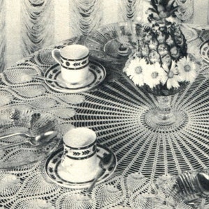 May include: A white crocheted tablecloth with a delicate lace pattern. Two white teacups with black and white designs are placed on the tablecloth, along with a silver spoon and a glass vase with a pineapple and white flowers.