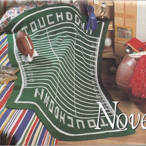 Può includere: Coperta a uncinetto verde che rappresenta un campo da football americano, con cuciture e linee di yard bianche. La coperta ha le parole "TOUCHDOWN" in alto e "YARDS TO GO" in basso.