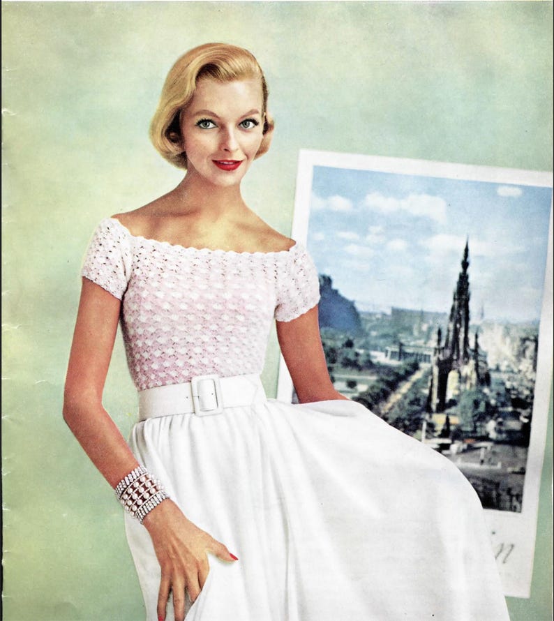 May include: A short-sleeved, off-the-shoulder top with a light pink, textured, scalloped knit pattern. The top is paired with a solid white, high-waisted skirt featuring a wide white belt with a rectangular buckle and a multi-strand beaded bracelet.