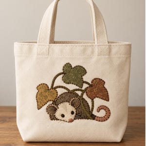 May include: A cream-colored canvas tote bag featuring a felt appliqué of an opossum nestled among autumn-colored leaves. The opossum is gray and white with a pink nose, and the leaves are shades of green, yellow, and brown with visible stitching.