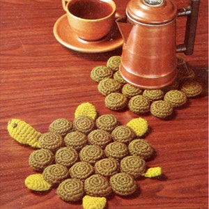 May include: Two crocheted pot holders, one shaped like a turtle and the other a circle, with brown and yellow yarn. The turtle pot holder has a brown body and yellow head, legs, and tail. The circular pot holder is made of brown yarn. A brown coffee pot and a brown cup and saucer are on a wooden table.