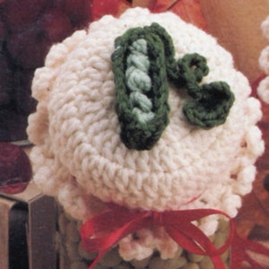 May include: A white crocheted jar lid with a green crocheted pea pod design. The lid is tied with a red ribbon.