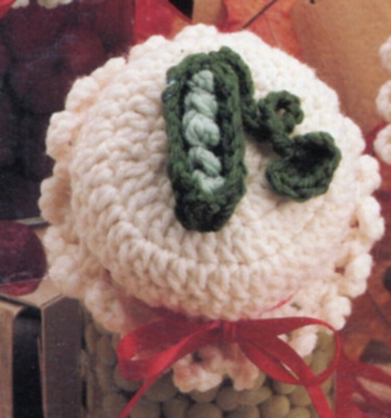 May include: A white crocheted jar lid with a green crocheted pea pod design. The lid is tied with a red ribbon.