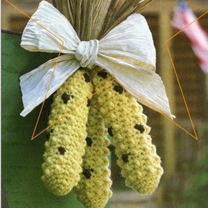 May include: Three crocheted corn cobs with brown spots, tied together with a white bow. The cobs are yellow and have a textured surface.