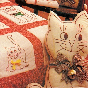 May include: A white plush cat pillow with a red and white striped bow and a gold bell. The pillow is sitting on a red and white quilt with embroidered cats.