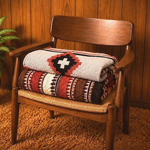 May include: Two folded woven blankets stacked on a wooden chair. The top blanket is light gray with a red, black, and white geometric diamond pattern. The bottom blanket features a dark brown, red, and cream striped pattern with textured details.