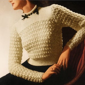 May include: Cream-colored textured knit sweater with long sleeves and a ribbed hem. The sweater features a round neckline with a black bow detail. The model is wearing black pants and sitting on a red chair.