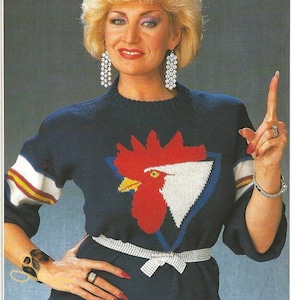 Könnte beinhalten: Eine Frau trägt einen blauen Pullover mit einem weißen und roten Hahn-Design. Sie trägt einen silbernen Gürtel und hat ihre Haare zu einem kurzen, blonden Bob gestylt. Sie zeigt mit dem Finger nach oben und lächelt.